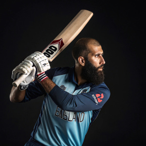 Cricket player Moeen Ali, wearing the England team jersey. Holding a bat in a batting pose. The dark background highlights the focus on him.