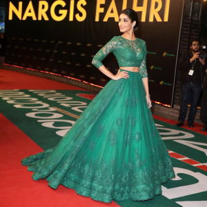 Nargis Fakhri on the set of a Bollywood film premiere. She wears a stunning emerald green gown with intricate lace detailing and a voluminous skirt. The red carpet is adorned with a bold green and black pattern, while promotional logos for the film loom large in the background.
