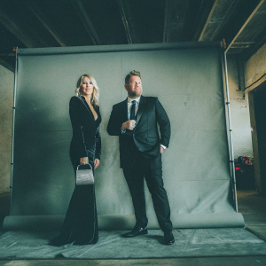 Julia Carey and James Corden at the Golden Globe Awards. Julia in a black velvet gown and clutch. James in a formal dark suit and tie. Both standing against a plain black backdrop.