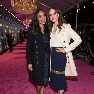 Rosario Dawson and Eliza Dushku at a glamorous awards show. Both in elegant attire, smiling for the cameras. Eliza in a black coat and skirt, heels, while Rosario wears a white coat with gold accents and navy skirt. Setting includes a chandelier and pink carpet.