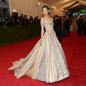 Fei Fei Sun at Met Gala. Wearing an elegant off-the-shoulder gown with vertical stripes in beige and gray. Standing on a red carpet with a crowd of elegantly dressed guests in the background.