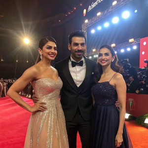 Jacqueline Fernandez, John Abraham, and Rakul Preet Singh at an award ceremony. Dressed in formal attire, posing together on stage with bright lights and cameras flashing.