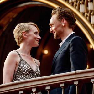 Mia Wasikowska and Tom Hiddleston at Cannes Film Festival premiere. Mia in black and white dress, Tom in dark navy suit. Friendly expressions. Dark backdrop with gold details