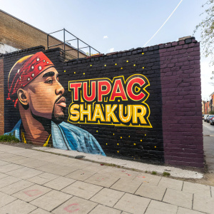 Tupac Shakur's portrait. Graffiti-style mural on a brick wall. Vibrant colors and bold text in a retro font. Urban setting, blue sky in background.
