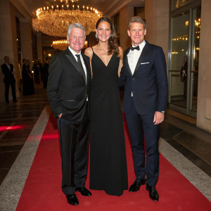 Marcel Reif, Ana Ivanovic-Schweinsteiger, and Bastian Schweinsteiger attend a gala evening. Ana, dressed in an elegant long black gown, stands between the two gentlemen. The backdrop showcases a sparkling chandelier and red carpet.