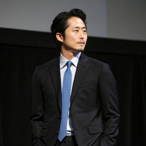 Stephen Yeun at the press junket. Black suit, wearing a blue tie. Simple dark background