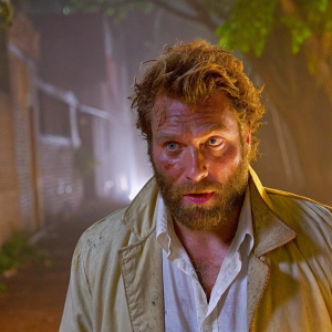 Jai Courtney, rugged look with scruffy beard and tousled hair. Light jacket over white shirt. Dark, blurred urban backdrop
