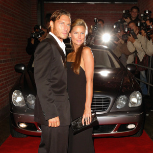 Francesco Totti and Ilary Blasi at a luxury car launch event. They are standing in front of a gleaming Mercedes-Benz S-Class car, posing for photographers on a vibrant red carpet.