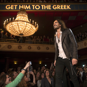 Russell Brand at a packed concert venue. He's standing under a chandelier with a banner that reads 'GET HIM TO THE GREEK'. Crowded with fans including a woman taking a photo.