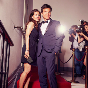 Jason Bateman and Amanda Anka attending a glamorous award ceremony. Jason in a tailored navy suit, Amanda in a sleek black dress. Flashing cameras, red carpet ambiance