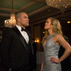 Tito Ortiz and Amber Nichole Miller at a glamorous gala. Tito, dressed in a black tuxedo, looks towards Amber, who wears a sparkling silver dress. Dimly lit room with chandeliers.