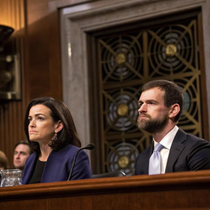 Sheryl Sandberg and Jack Dorsey in formal suits during a congressional hearing. Ornate details and intricate ironwork visible in the background.