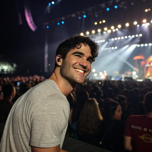 Darren Criss, wearing a casual grey t-shirt, smiles at the camera. Behind him, a crowded concert venue with live music