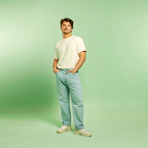Gregg Sulkin, actor, in casual wear. White t-shirt and jeans. Grey backdrop. Poses confidently with hands in pockets. Professional photo shoot.