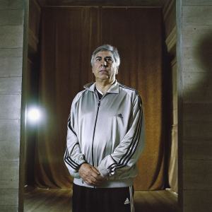 Professional photograph of Carlo Ancelotti. Older man with graying hair and serious expression. Wears Adidas tracksuit with logo. Black tracksuit pants. Composed stance. Solid blue backdrop