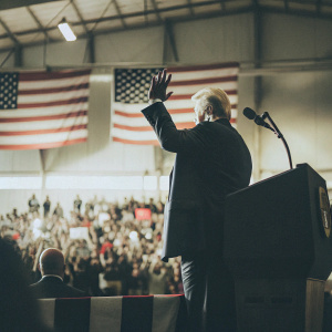 Donald Trump at a bustling campaign rally, waving from the podium. Behind him, a sea of supporters with American flags waving.