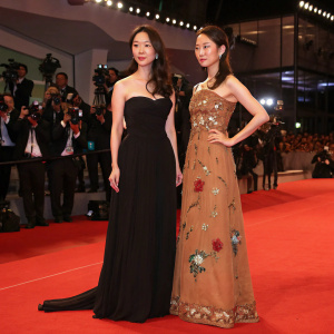 Actresses Tang Wei and Lee Yeon Hee at a glamorous fashion event. Both wear elegant gowns. Tang Wei in black, Lee Yeon Hee in brown with embroidery. The setting is an indoor red carpet event with flashing cameras and a crowd.