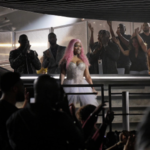 Nicki Minaj, with vibrant pink hair and glitzy dress, center stage at an awards show. Crowd in the background