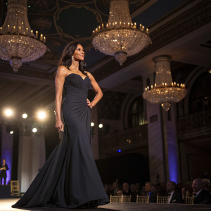 Padma Lakshmi, in elegant evening gown, hosting event at grand ballroom. Dim lighting and chandeliers in background.