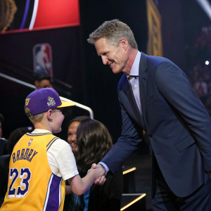 Steve Kerr, dressed in a suit, shaking hands with a fan at an NBA awards ceremony. The fan is wearing a Lakers jersey.