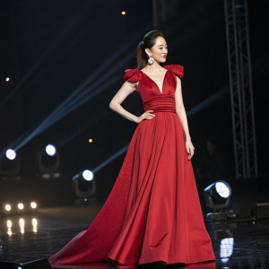 Dilraba Dilmurat at a glittering awards night. Wearing an elegant red dress with voluminous top and flowing skirt. Dark stage with spotlights