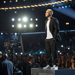 Eminem on a podium at award show. Black suit and white shirt. Behind him, a sea of flashing cameras and a bustling crowd.