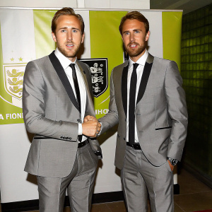 Formal portrait of Harry Kane and Gareth Southgate. Wearing matching gray suits, shaking hands. Sponsored event backdrop.