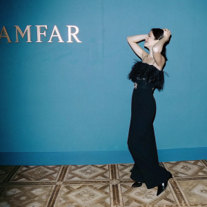 Alessandra Mastronardi, wearing a black floor-length gown with intricate fringed bodice, posing elegantly. Event backdrop with amfAR logo