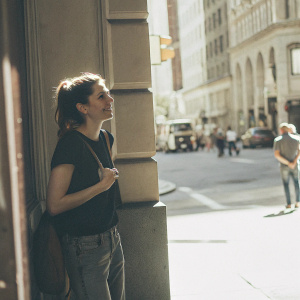 Daisy Ridley, standing against a busy city street backdrop. She wears a casual outfit, black t-shirt and jeans. Hair styled in a ponytail. Focus on her casual, relaxed expression.