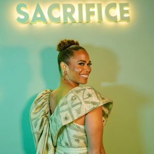 Paula Patton at the BET+ series 'Sacrifice' premiere. Dark blue backdrop with glowing 'Sacrifice' title. She wears a high-waisted, off-the-shoulder patterned dress with gold accents, smiling confidently.