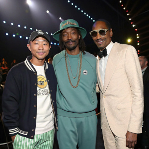 Pharrell Williams, Snoop Dogg, and a person of Asian descent stand together at the Grammy Awards ceremony. Casual outfits, smiling