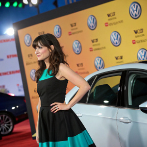 Zooey Deschanel at a car unveiling event, standing confidently in a black and teal dress. Vibrant backdrop featuring Volkswagen logos and branding