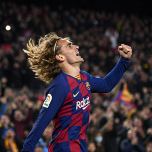 Antoine Griezmann, with wild blonde hair, celebrating a goal. The Barcelona FC jersey stands out with its iconic horizontal stripes. The background is filled with cheering fans at Camp Nou stadium.