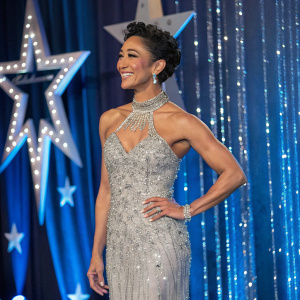 Karen Hauer at a gala event, dressed in a sparkling silver gown with intricate beadwork. She is standing confidently with a radiant smile. The backdrop is a shimmering blue curtain with embedded stars.
