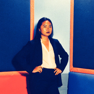 Professional headshot of a young Asian woman, wearing a black blazer over a white blouse. Confident pose with one hand on hip. Brightly lit studio setting