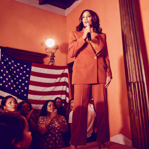 Kamala Harris, wearing a red power suit, addressing a crowd at a rally. Bright stage lights, patriotic backdrop.