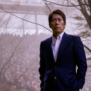 Akira Nishino, serious face, in sharp navy suit. Blurry crowd in background.