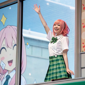 Illustration of a cheerful young anime girl with fair skin and pink hair, standing in front of a window. Wearing a white shirt and a green plaid skirt, waving happily. The scene is set against a digital backdrop with cartoonish elements.