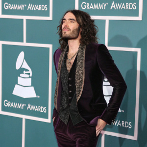 Russell Brand at the Grammy Awards. Wearing a dark velvet suit with layered vest, teal backdrop with Grammy logo