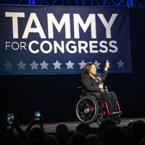 Tammy Duckworth, seated in wheelchair, addresses crowd at rally. Dark stage background with spotlight on her. Banner reads 'Tammy for Congress'.