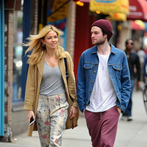 Sienna Miller and Tom Sturridge strolling down a vibrant street. Miller's blonde locks flow freely, donning a tan jacket over a gray top with floral-patterned pants. Sturridge sports a white shirt, blue denim jacket, and maroon beanie.