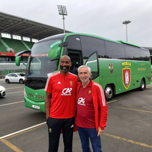 Nicolas Anelka and Raymond Domenech, both in red team jerseys, stand near a green sports bus with team logos. Stadium parking lot