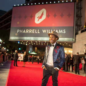 Pharrell Williams, standing confidently on a bustling red carpet at a major film festival. Behind him, a large screen displays the event's logo and sponsors.