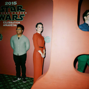 At the 2015 Star Wars Celebration in Anaheim, John Boyega, Daisy Ridley, and J.J. Abrams pose for a picture. The backdrop is filled with Star Wars logos and text. John Boyega stands left, Daisy Ridley is centered, and J.J. Abrams, wearing glasses, is on the right.
