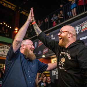 Brian Posehn and Jamey Jasta, both balding men with beards, high-fiving joyfully at a concert. Sponsor logos on the banner behind them.