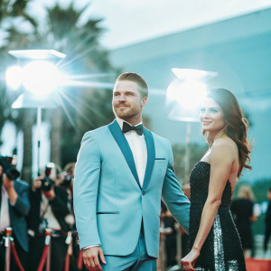 Stephen Amell and Nina Dobrev at the Critics' Choice Awards. Stephen in a sleek blue tuxedo, Nina in a shimmering black gown with silver details. They are posing on the red carpet surrounded by flashing cameras.