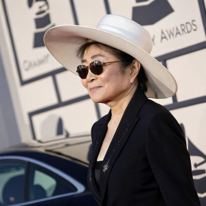 Yoko Ono, Asian female with mature appearance, wearing a wide-brimmed white hat and dark jacket. She poses with a slight smile at the Grammy Awards. Patterned backdrop with car partially visible in background. High resolution photo.