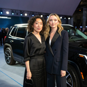 Sandra Oh and Jodie Comer at a car launch event. Both standing in front of a sleek black SUV. Neutral expressions, formal attire.