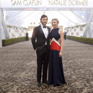 Sam Claflin and Natalie Dormer at a gala event. Formal attire. Black and white tuxedo and color-blocked dress. Backdrop with event logos.