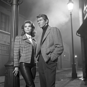 Jennifer O'Neill and James Coburn, stars of 'The Carey Treatment', in a vintage Hollywood scene. Jennifer in a gingham jacket with leather pants, James in a tailored blazer, both in a black and white setting.